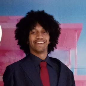 Retrato de homem jovem sorrindo, com cabelo black power escuro, usando terno azul-marinho e gravata vermelha.