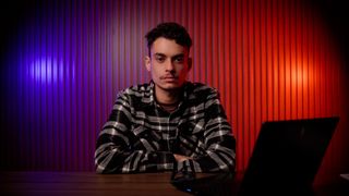 Homem jovem de camisa xadrez, sentado à mesa com os braços cruzados ao lado de um notebook, diante de uma parede ripada iluminada de vermelho e roxo.